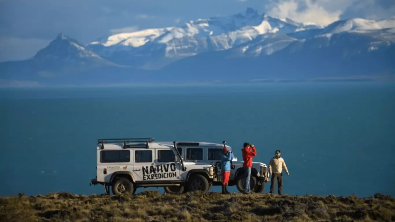 Travellers beside 4x4 vehicles at a clifftop lookout over Lago Argentino with the snow-capped Andes behind