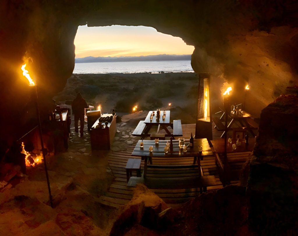 Tables set inside a sandstone cave with torches burning and the arch framing Lago Argentino at sunset