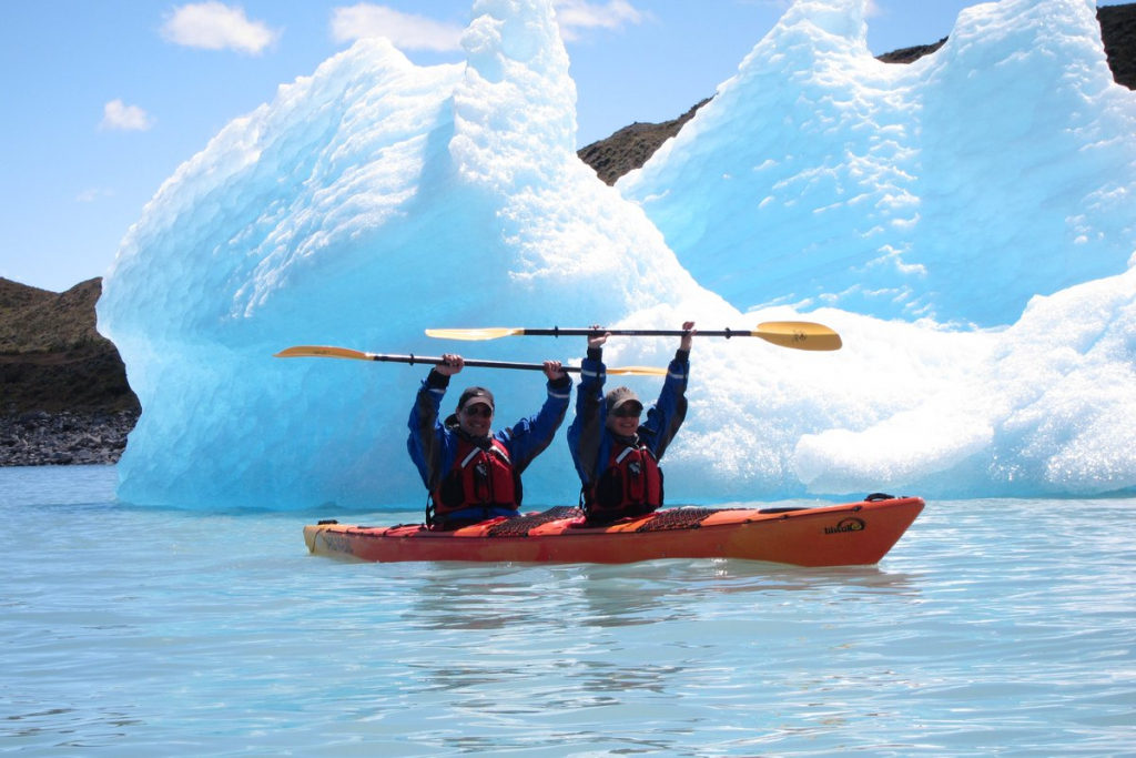 Two kayakers raising their paddles beside a large blue iceberg on Lago Argentino